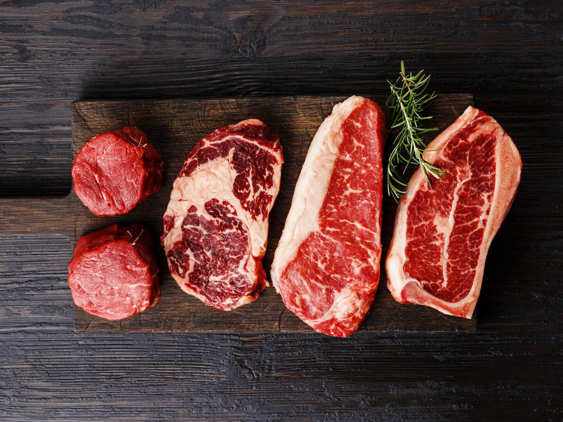 AdobeStock_193571214 Assorted cuts of premium beef, including tenderloin and ribeye, displayed on a wooden cutting board with a sprig of rosemary for garnish. Ideal for gourmet cooking and grilling enthusiasts.