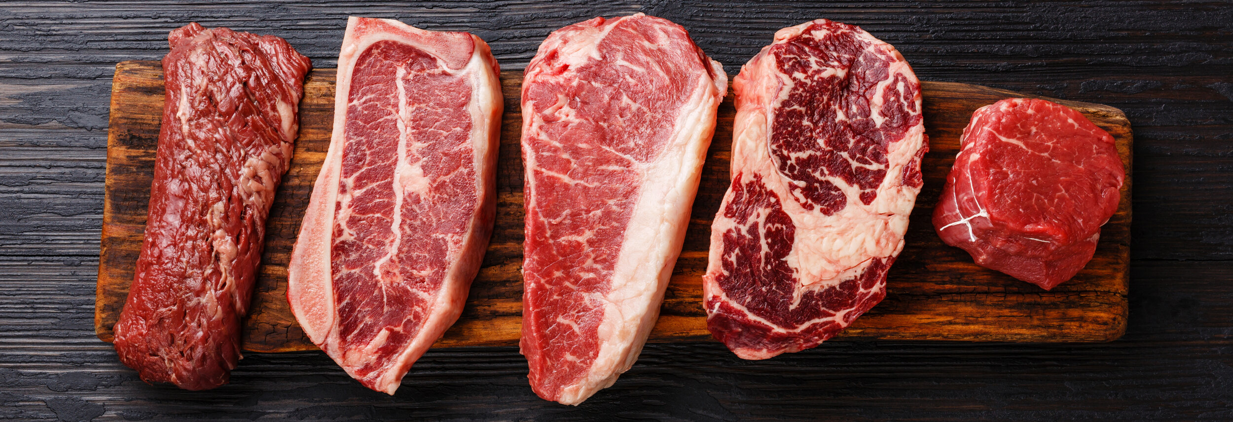 Variety of Raw Black Angus Prime meat steaks Machete, Blade on bone, Striploin, Rib eye, Tenderloin fillet mignon on wooden board Assorted cuts of fresh beef displayed on a wooden cutting board, including ribeye, sirloin, and tenderloin, showcasing marbling and texture ideal for grilling or roasting.