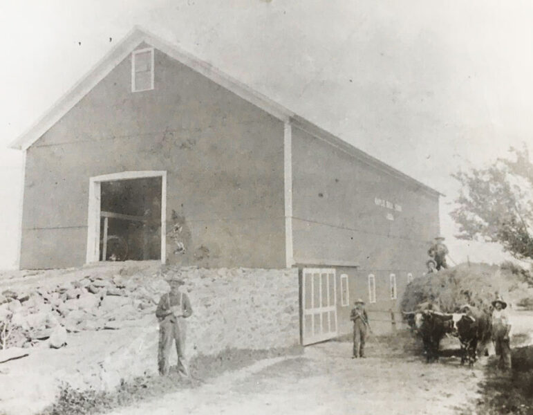 Historic black and white photo of a barn with men and oxen outside, showcasing early agricultural practices and rural life in the past. The barn features a stone foundation and a large entrance, with individuals engaged in farming activities.
