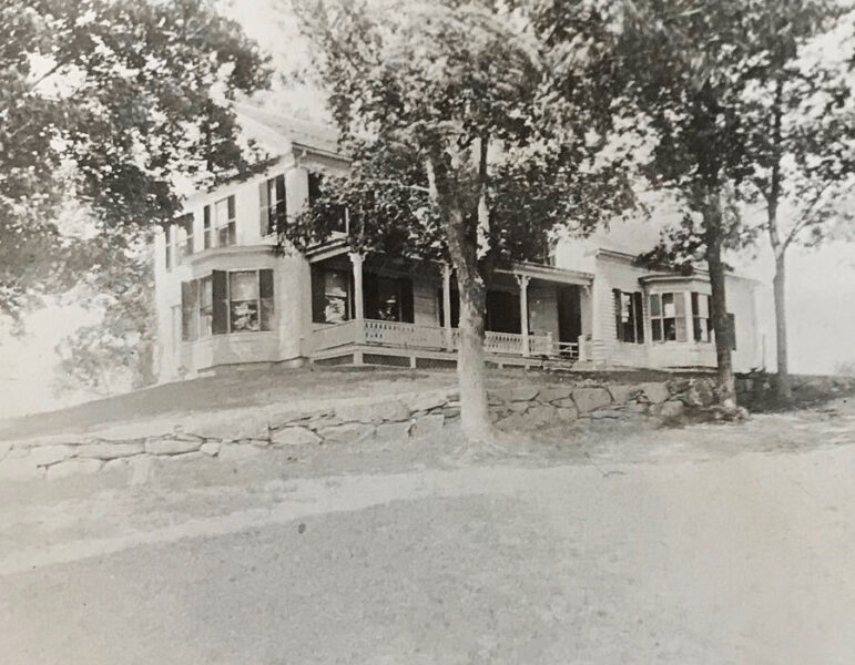 Historic two-story house surrounded by trees, featuring a large front porch and multiple windows, captured in a vintage black-and-white photograph. The home is situated on a slight hill with a stone wall in the foreground, showcasing classic architectural details.