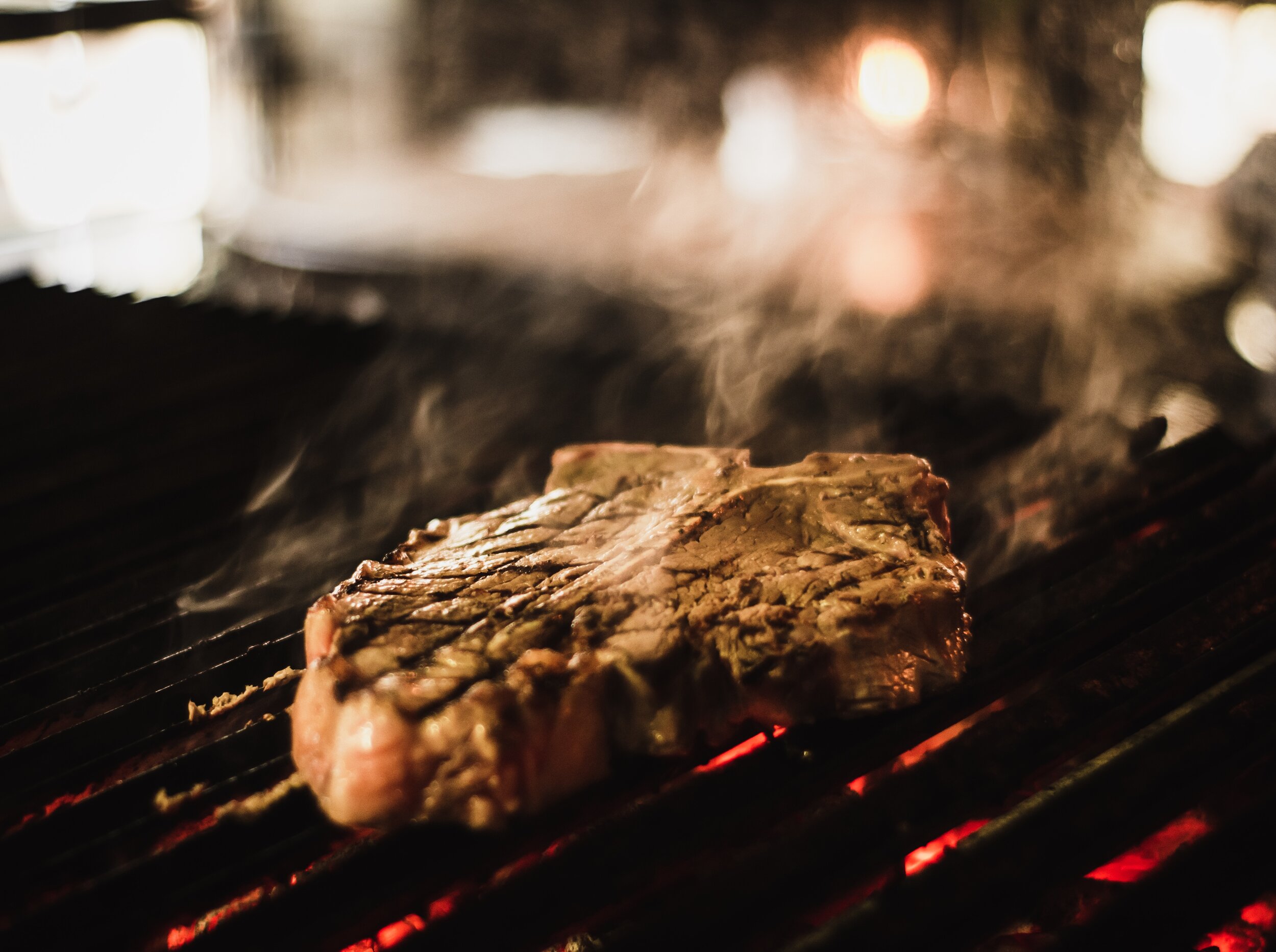Grilled steak sizzling on a barbecue grill, emitting steam and smoke, with visible grill marks and glowing coals in the background, showcasing the cooking process and rich textures of the meat.
